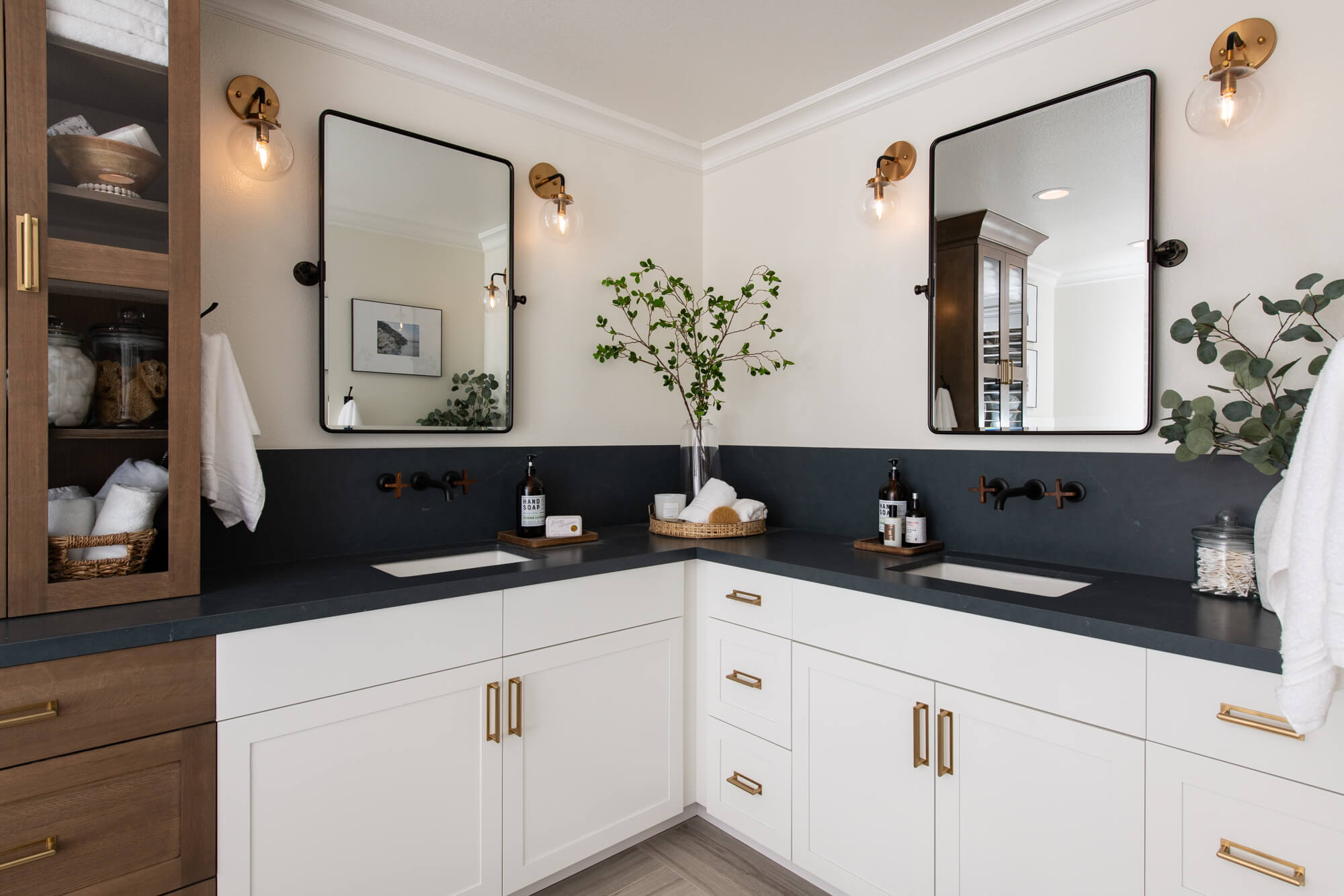 Kitchen and Bathroom Cabinets in a modern bathroom—white shaker vanity in a corner layout, dark quartz countertop/backsplash, brass pulls, wall-mounted faucets, wood storage tower, and sconce lighting.
