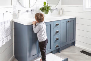 Kitchen and Bathroom Cabinets: blue shaker bathroom vanity with white quartz top, dual sinks, wall-mount faucets, subway tile, and a pull-out step for a child.