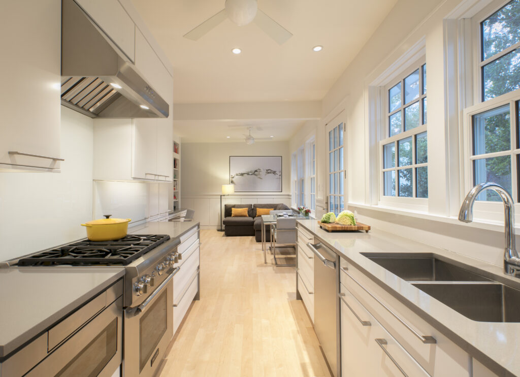 Contemporary galley kitchen cabinets with glossy white finish, stainless steel appliances, grey countertops, and corridor layout connected to an open living room