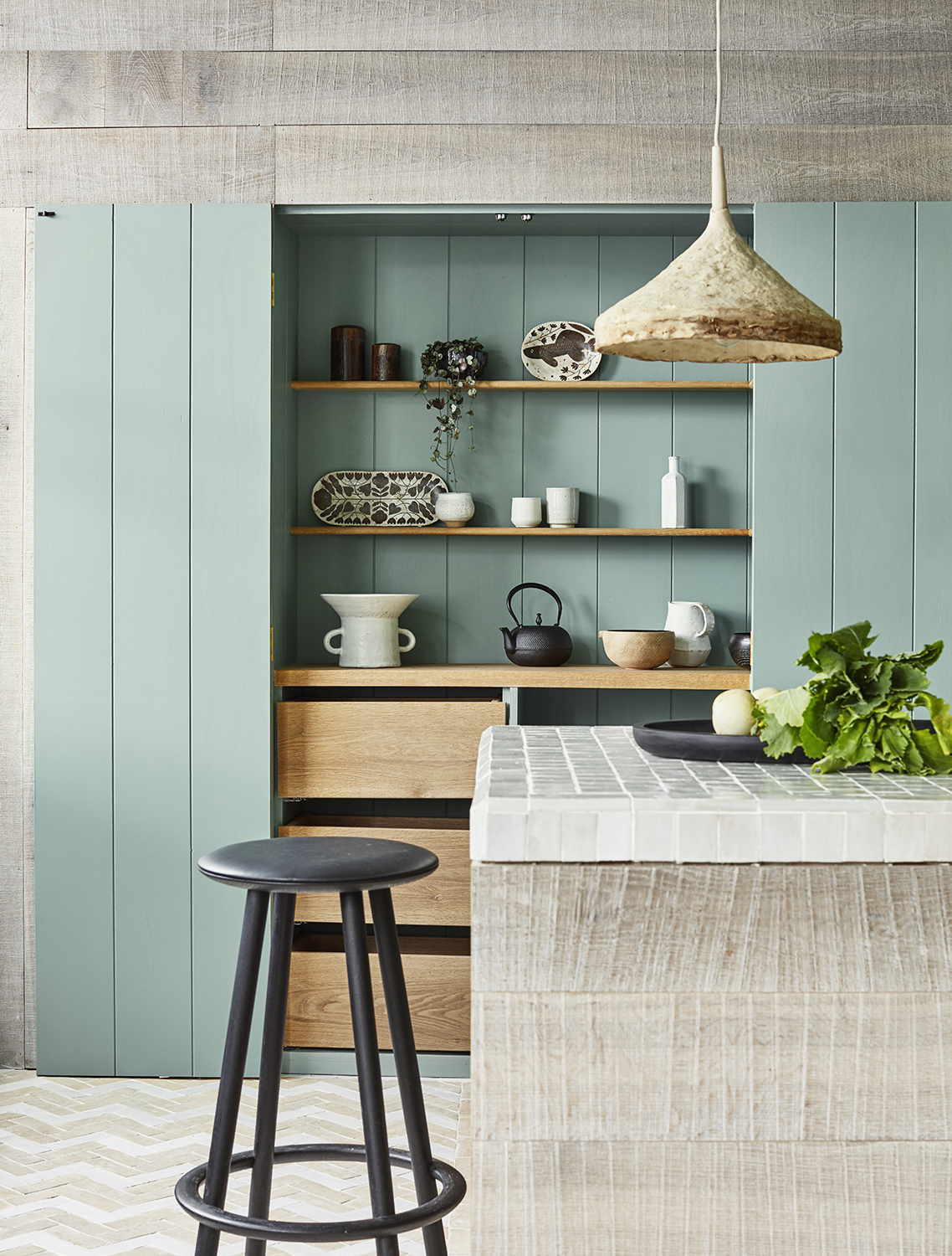 Kitchens and Bathrooms: minimalist sage-green pantry nook with open wood shelves, handmade-tile island, rustic pendant light, and black bar stool.