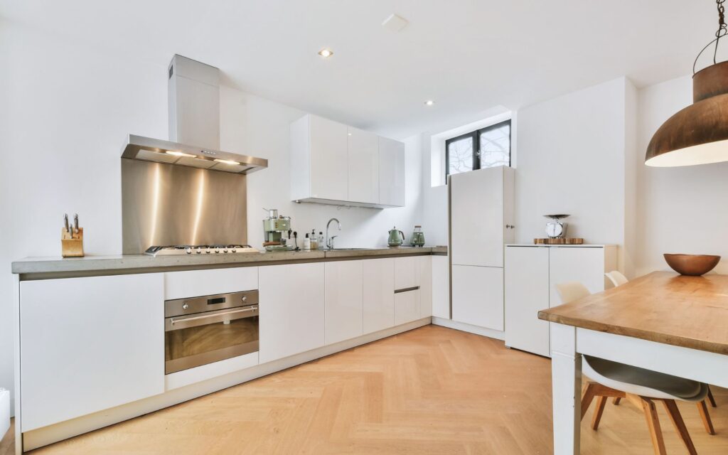 Bright modern L-shaped kitchen cabinets for rental properties with glossy white flat-panel doors, stainless range hood and backsplash, integrated oven, and light wood herringbone floor