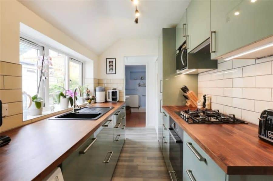 Narrow galley kitchen cabinets with sage green finish, wooden countertops, subway tile backsplash, and large windows bringing natural light into the corridor layout