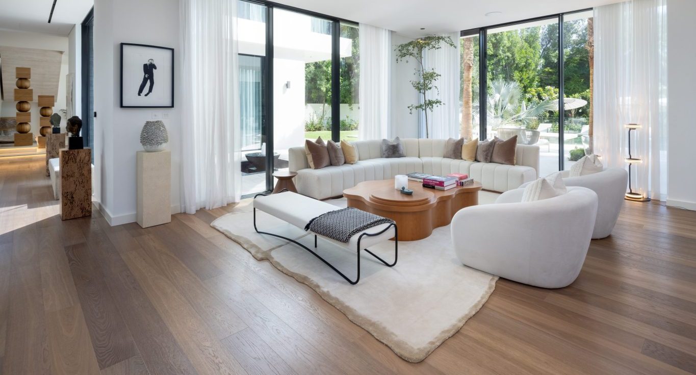 Premium RTA cabinets Canada: airy modern living room with a curved white sectional, rounded lounge chairs, organic wood coffee tables, bench on a plush rug, floor-to-ceiling windows, and wide-plank wood floors.