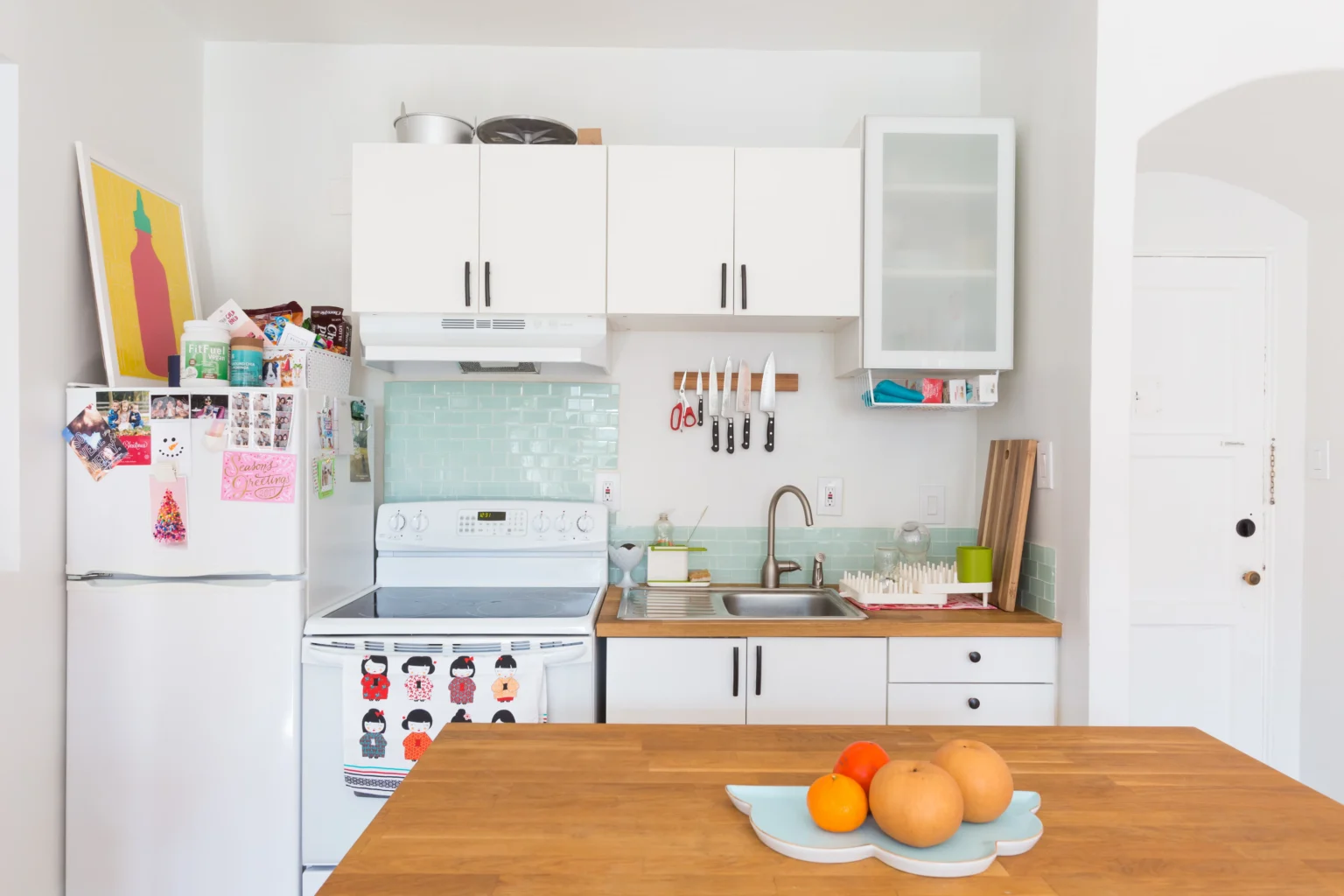 Small, bright apartment kitchen with white flat-panel RTA cabinets, wood countertops, mint-green subway tile backsplash, white appliances, and a butcher-block island with fruit.