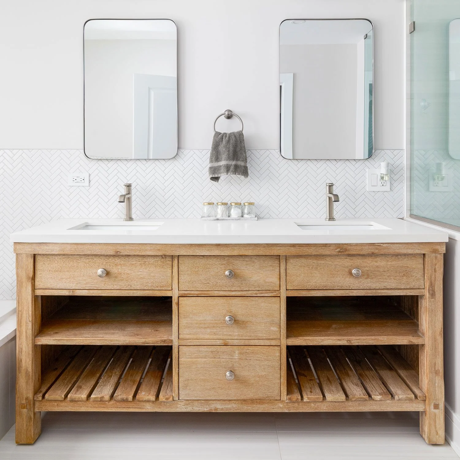 Rustic light-wood bathroom cabinets with open slatted shelves, center drawers, white quartz double-sink vanity, herringbone tile backsplash, and twin mirrors.