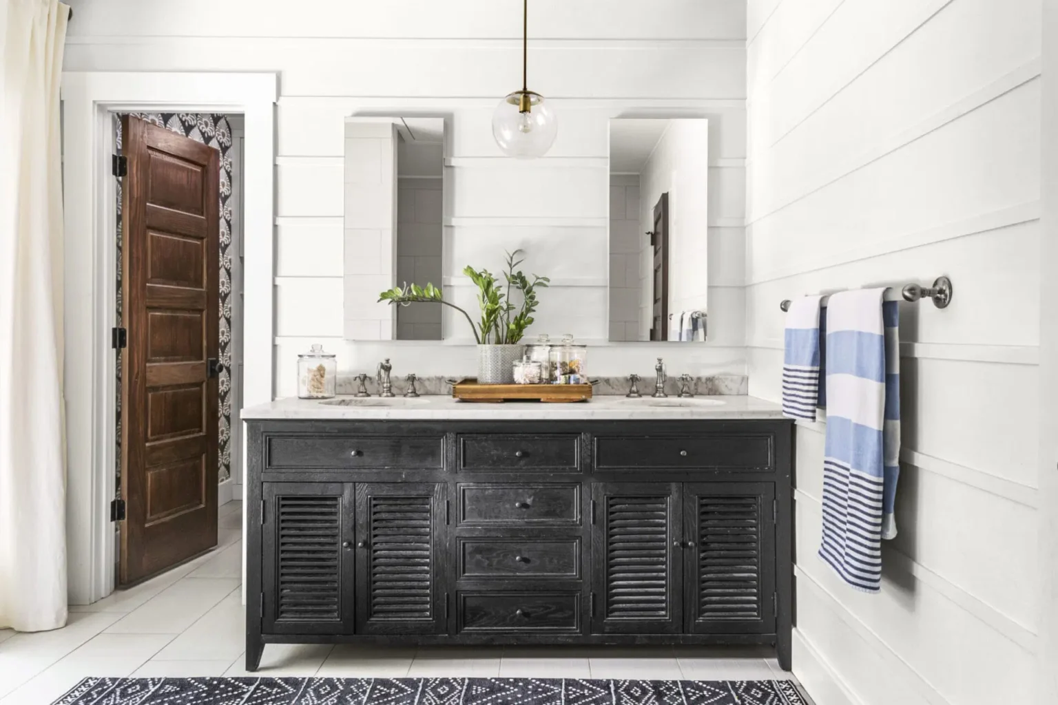 Bathroom with black double-sink vanity cabinets, marble countertop, twin mirrors, modern pendant light, and striped towels on a white paneled wall.