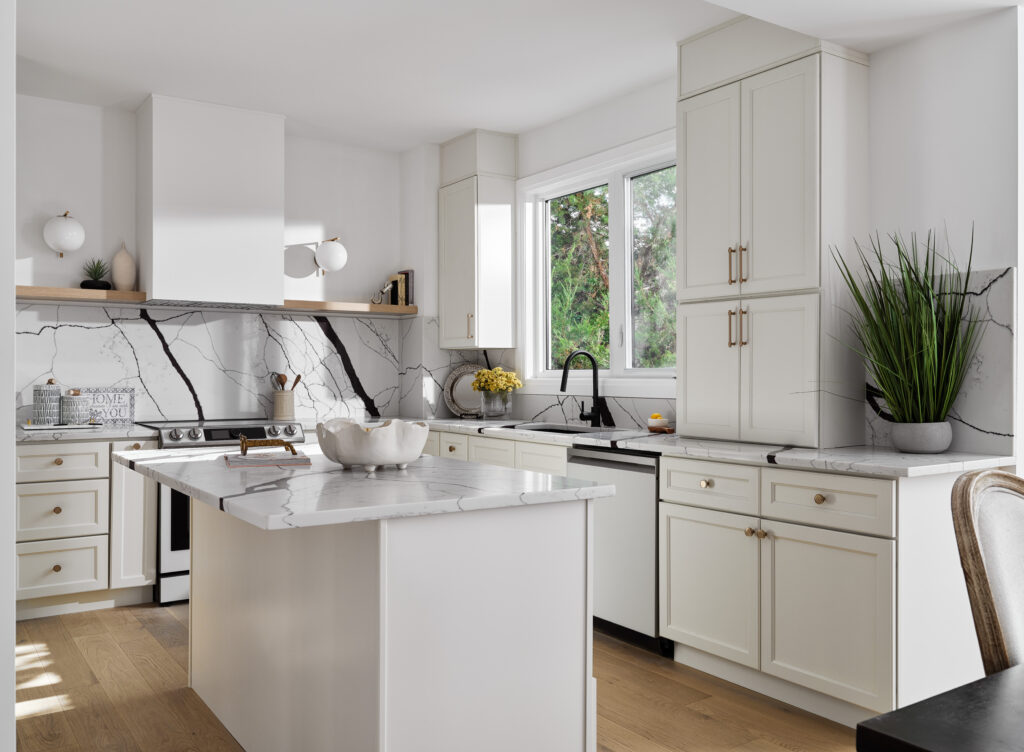 Bright kitchen with off-white shaker RTA cabinets, veined quartz waterfall island and slab backsplash, black faucet, brass hardware, floating shelf, and warm oak floors.