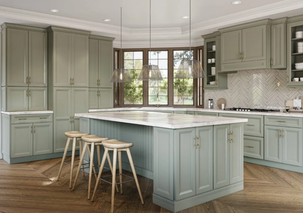Spacious kitchen with sage-green RTA cabinets, large beadboard island with marble-look countertops, brass hardware, herringbone backsplash, and herringbone wood floors under pendant lights.