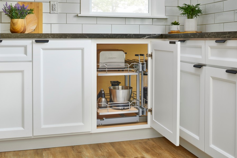 Close-up of white shaker RTA cabinets with black pulls; an open base cabinet reveals a pull-out organizer holding small appliances and bowls beneath a dark countertop and subway-tile backsplash.