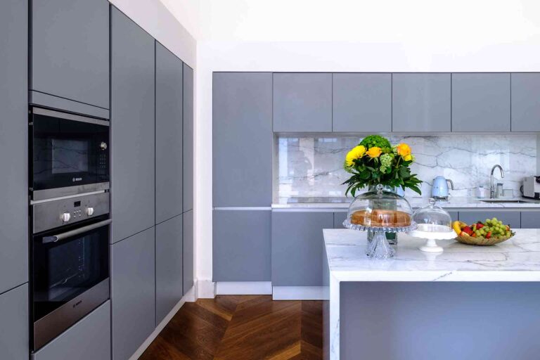 Sleek modern kitchen with gray flat-panel RTA cabinets, marble backsplash and waterfall island, built-in Bosch ovens, and herringbone wood flooring.