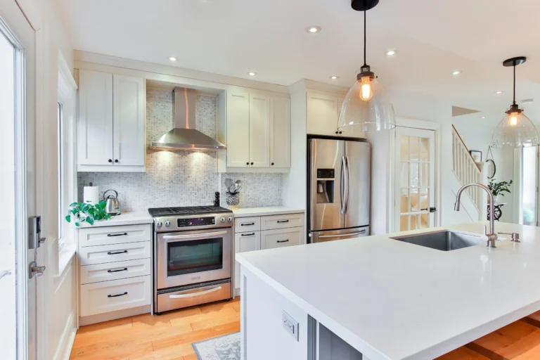 Bright kitchen with white shaker RTA cabinets, stainless appliances, mosaic tile backsplash, and a large quartz island with sink and glass pendants.
