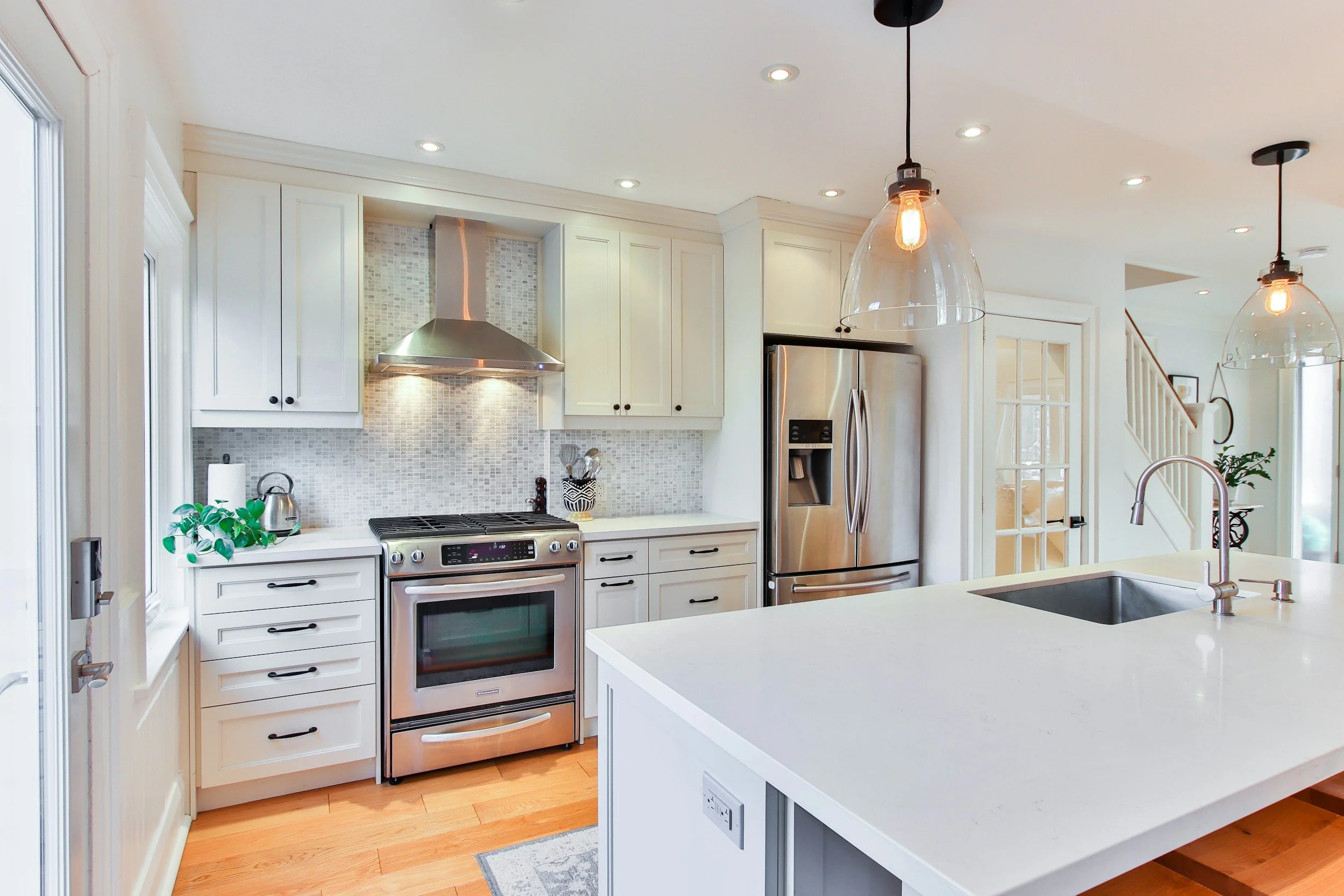 Bright kitchen with white shaker RTA cabinets, stainless appliances, mosaic tile backsplash, and a large quartz island with sink and glass pendants.