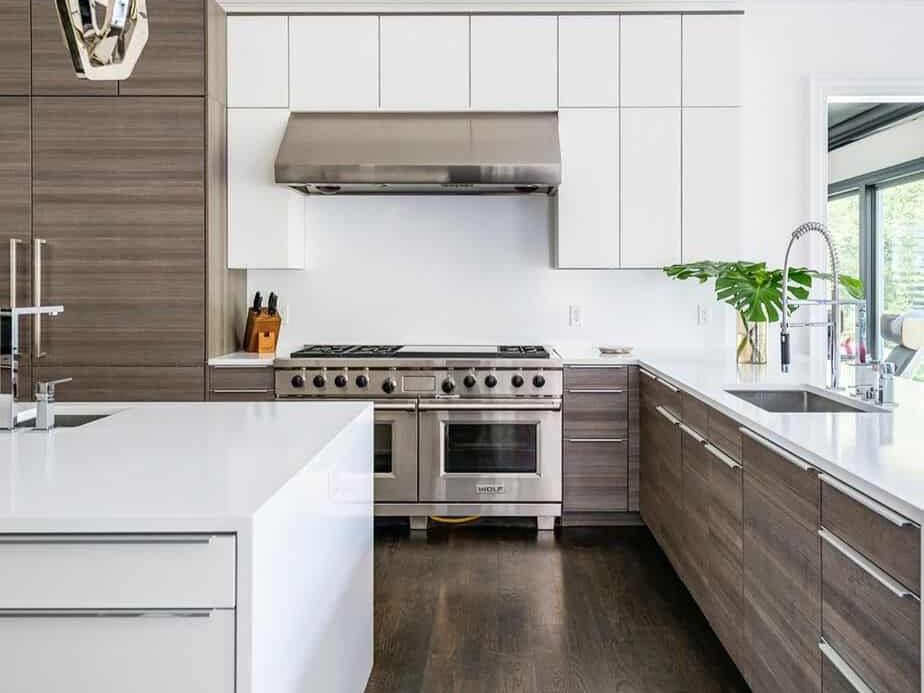 Sleek contemporary kitchen with wood-grain RTA flat-panel base cabinets, white slab uppers, quartz waterfall island, and a stainless pro range under a wide hood.