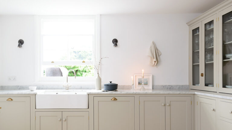 Bright classic kitchen with greige shaker premium RTA cabinets Canada, apron-front farmhouse sink under a window, marble-look countertops, glass-front hutch, and brass hardware.