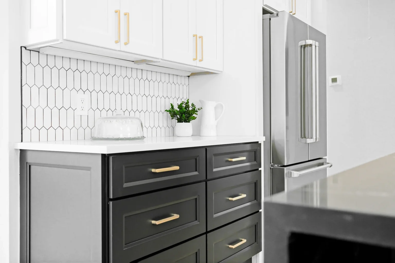 Close-up of a modern kitchen with white shaker Ready-to-Assemble (RTA) upper cabinets, charcoal shaker RTA drawer base, brass pulls, white quartz countertop, elongated hex tile backsplash, and a stainless French-door fridge.