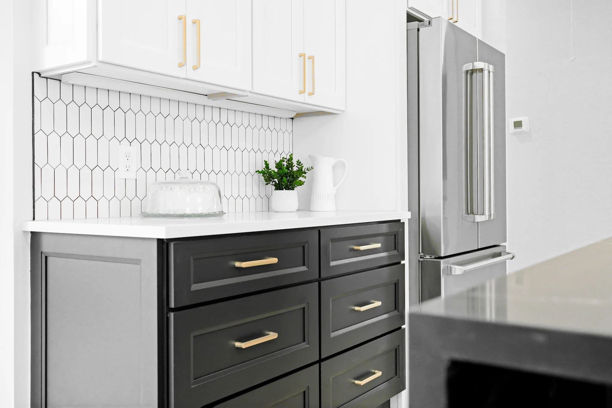 Close-up of a modern kitchen with white shaker Ready-to-Assemble (RTA) upper cabinets, charcoal shaker RTA drawer base, brass pulls, white quartz countertop, elongated hex tile backsplash, and a stainless French-door fridge.