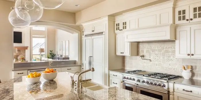Elegant traditional kitchen with cream RTA cabinets, raised-panel doors, glass-front uppers, granite countertops, patterned tile backsplash, paneled fridge, and an island sink beneath globe pendants.