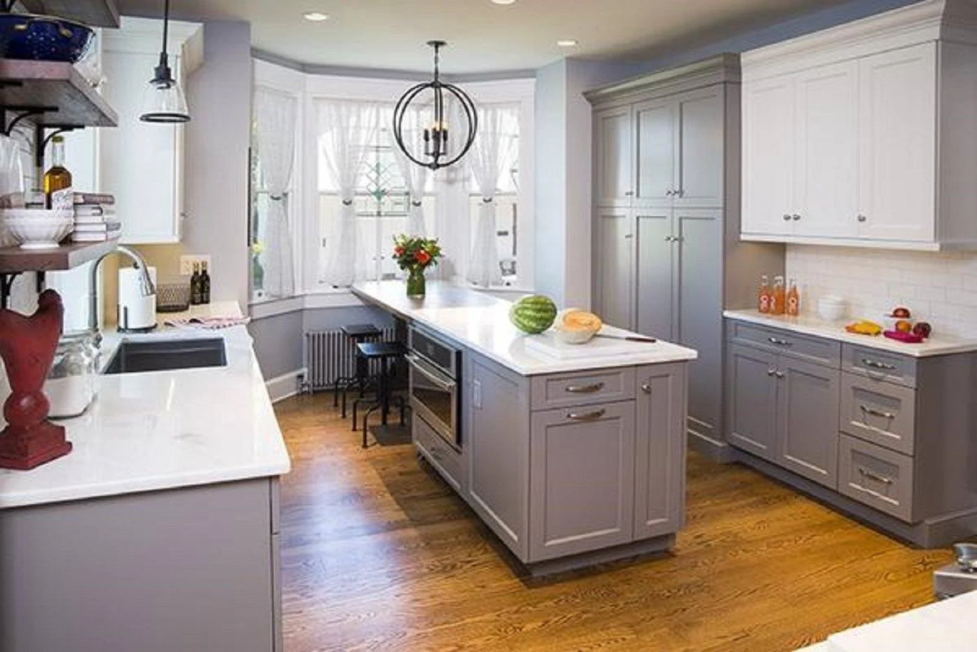 Two-tone shaker kitchen cabinet design in gray and white with a long quartz-topped island, built-in microwave, bay window, orb pendant, and warm wood floors.