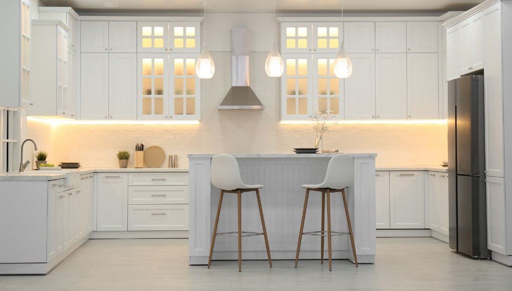 All-white kitchen cabinet design with glass-front uppers, warm undercabinet lighting, a petite island with two stools, stainless hood, and black fridge.