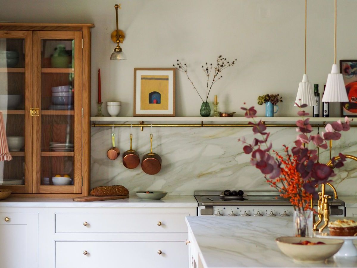 Cozy eclectic kitchen with white shaker kitchen cabinet drawers, an oak glass-front hutch, marble-veined backsplash and counters, brass rail with copper pans, and fluted white pendants.