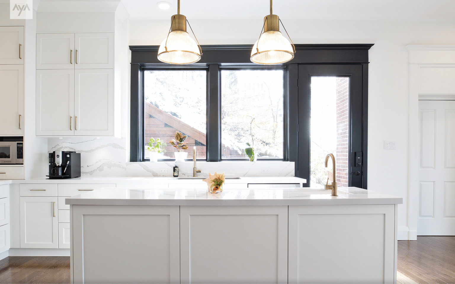 Bright white shaker kitchen cabinet with brass hardware, quartz-veined backsplash, large island, black-trimmed windows, and brass fixtures under glass pendants.