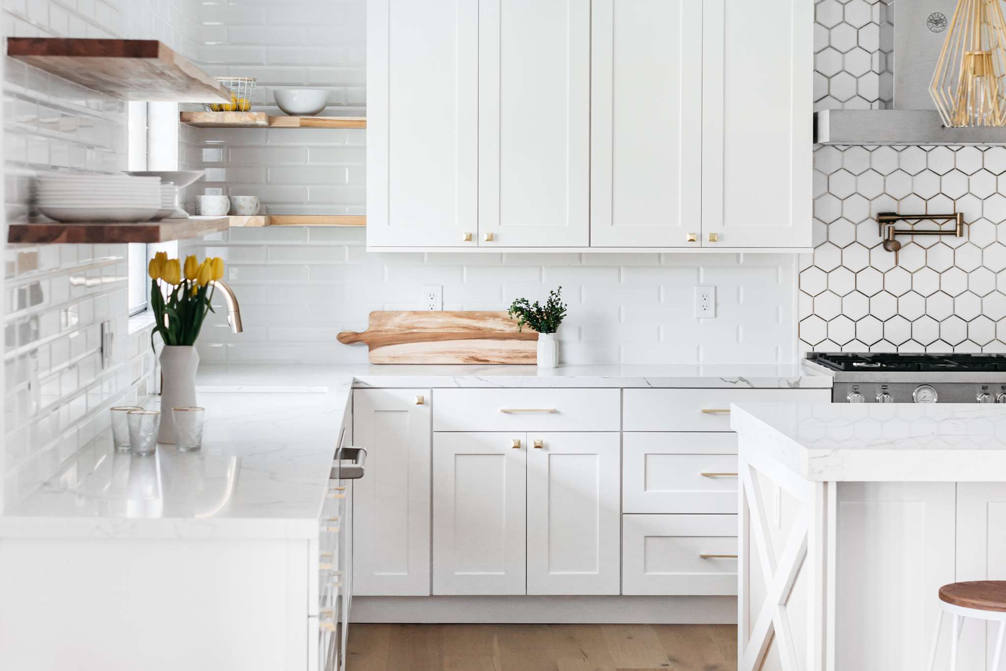 Bright white kitchen with shaker-style kitchen cabinet fronts and brass hardware, marble-look counters, floating wood shelves, mixed subway/hex tile backsplash, and an island with X-panel detail.
