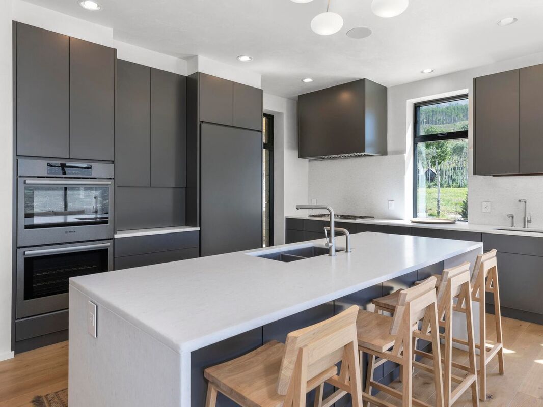 Modern matte-gray kitchen cabinets with an integrated fridge, large island with sink, and wood bar stools in a bright Canadian home