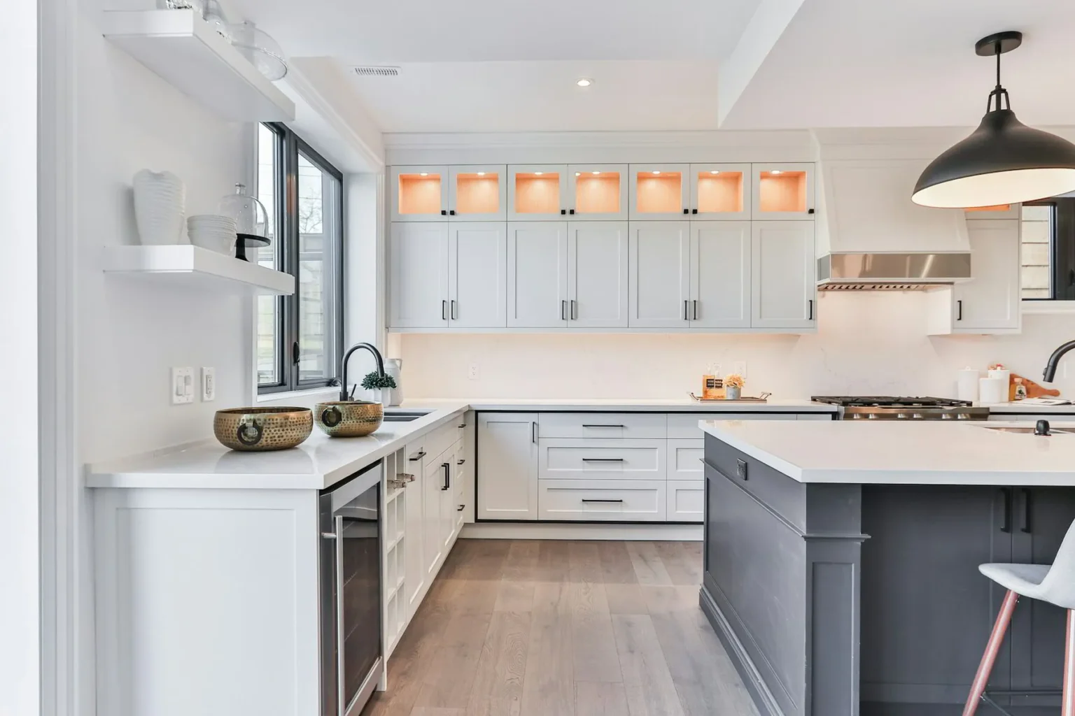 Contemporary kitchen with light gray shaker kitchen cabinet run featuring lit display uppers, white quartz countertops, stainless range hood, and a charcoal island under a black pendant.