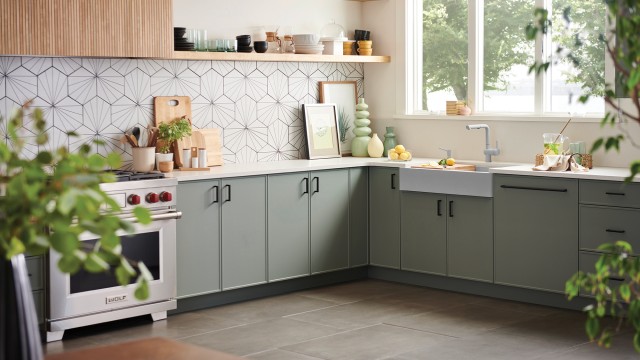 A modern L-shaped kitchen featuring sage green flat-panel kitchen cabinet units, black hardware, a white farmhouse sink, and a unique geometric tile backsplash.
