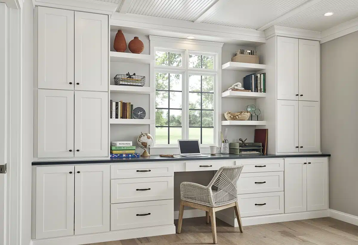 A built-in home office workstation created with white shaker kitchen cabinet units, featuring tall pantry cabinets, base drawers, a dark desk countertop, and open shelving around a window.