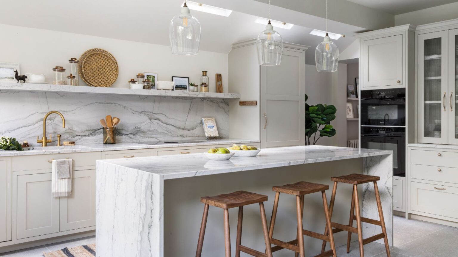 Light gray shaker kitchen cabinet run with marble backsplash and waterfall island, brass faucet and pulls, glass pendants, and wood stools in a bright modern space.