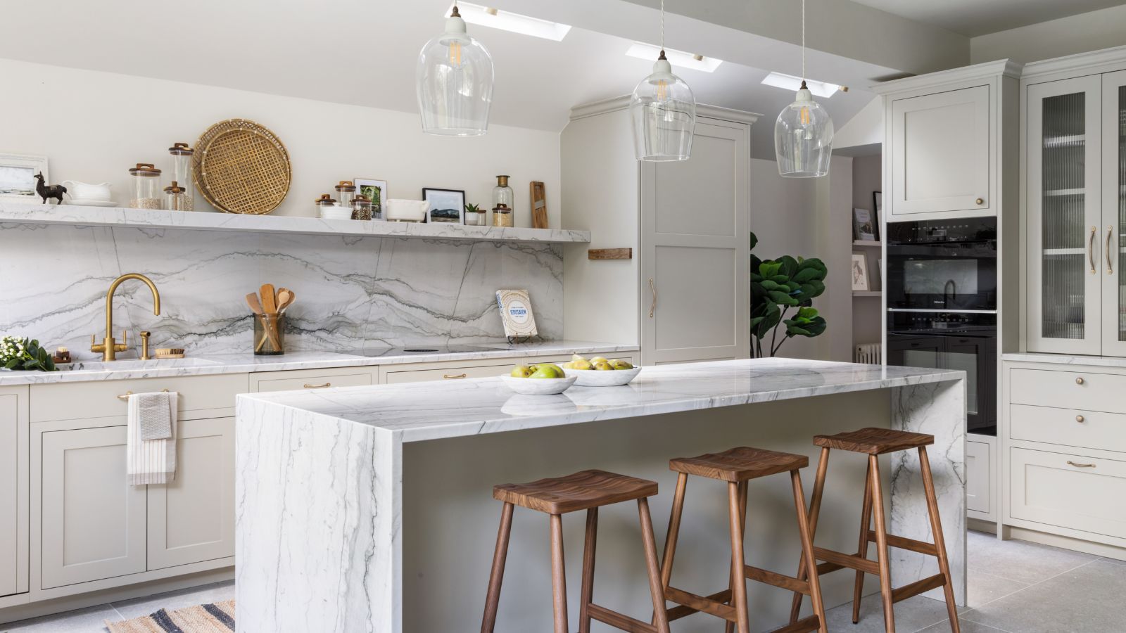 Light gray shaker kitchen cabinet run with marble backsplash and waterfall island, brass faucet and pulls, glass pendants, and wood stools in a bright modern space.