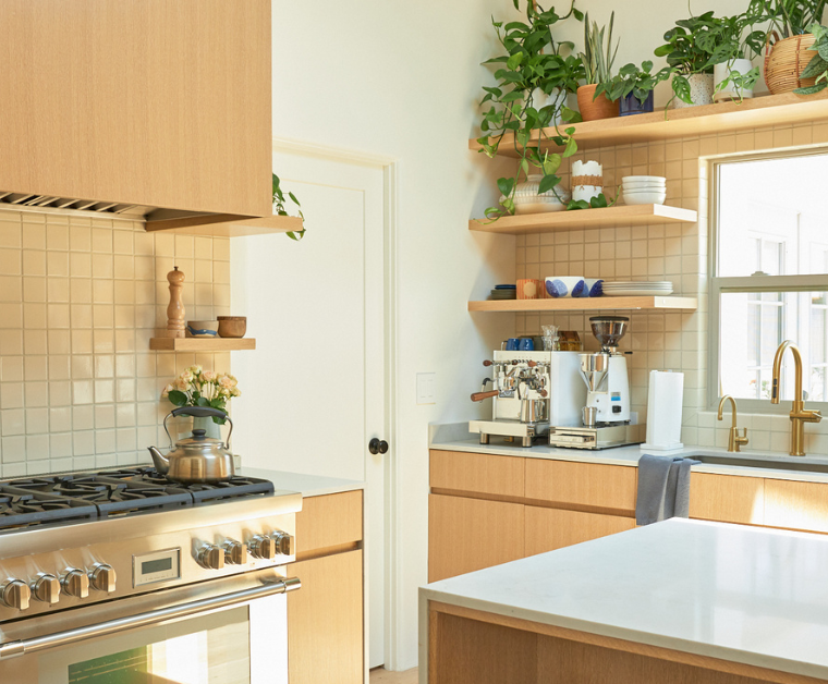 Bright modern kitchen with light wood kitchen cabinets, white countertops, open shelves filled with plants, and a coffee station by the window.