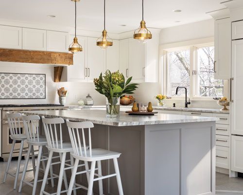 Sunlit modern farmhouse kitchen with white shaker kitchen cabinets, soft-gray island, marble-look countertops, brass pendants, patterned tile backsplash, and a rustic wood hood beam.