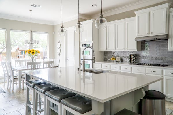 Bright modern white kitchen with shaker cabinets, large quartz island, pendant lights, and stainless steel range hood from kitchen suppliers.
