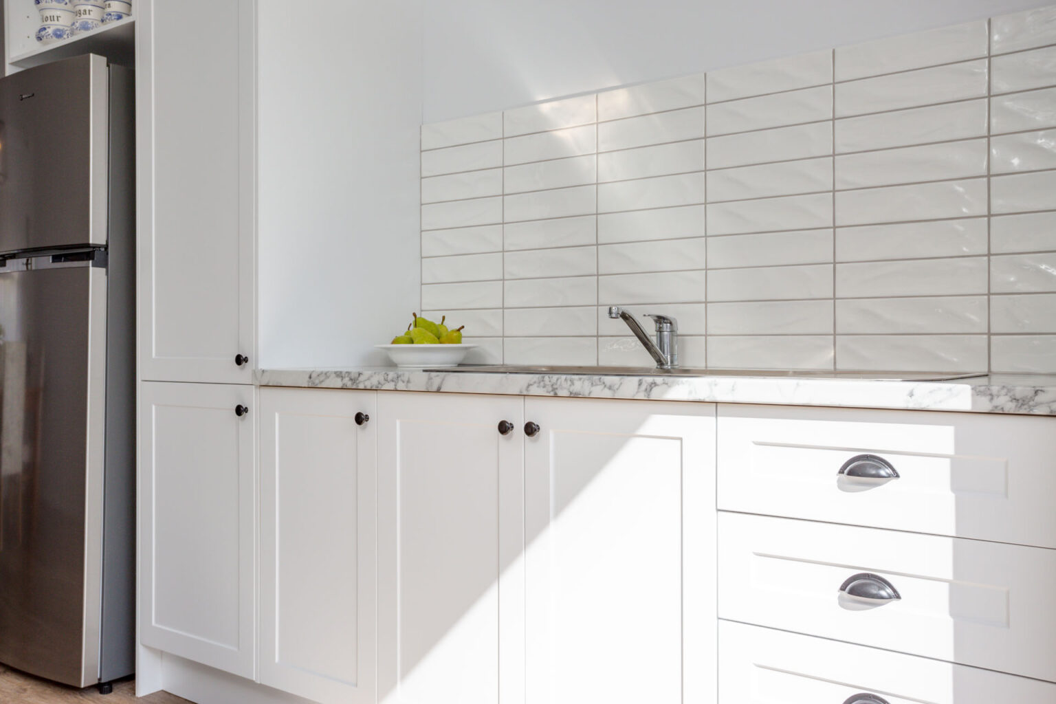 Bright white flat pack kitchen cabinet run with shaker-style doors, marble-look countertop, stainless faucet, and glossy subway tile backsplash beside a fridge.