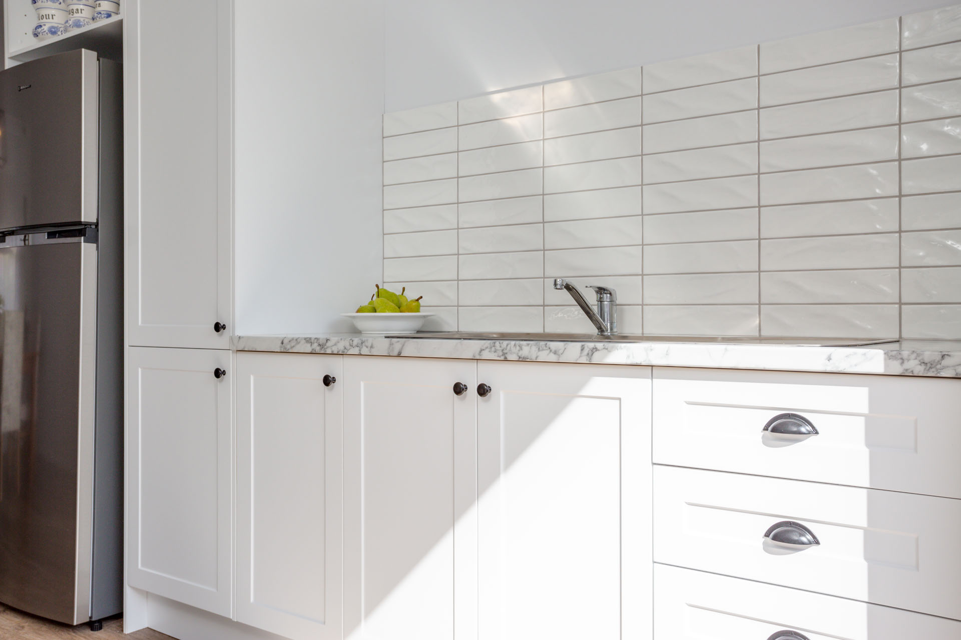 Bright white flat pack kitchen cabinet run with shaker-style doors, marble-look countertop, stainless faucet, and glossy subway tile backsplash beside a fridge.