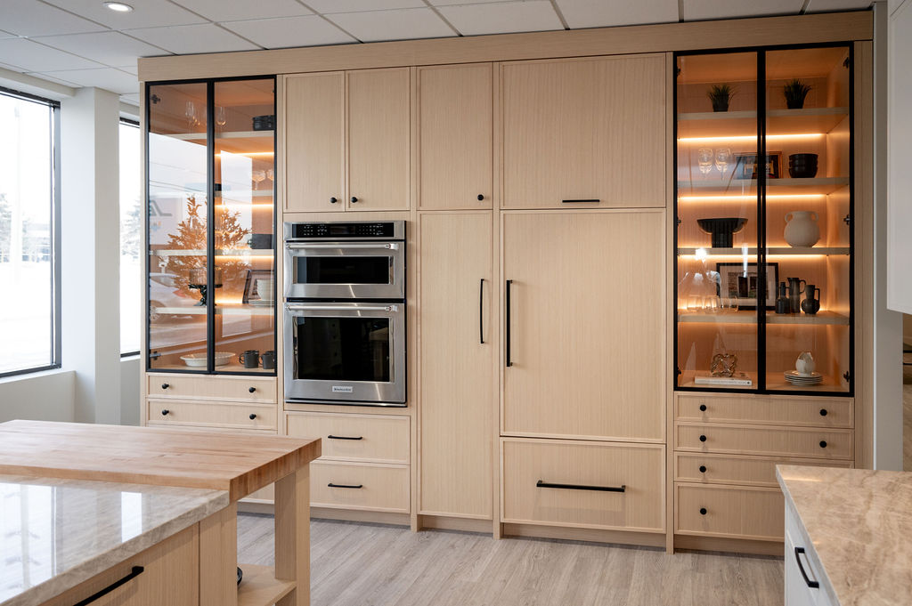 Modern light-wood Kitchen cabinet wall with built-in double oven, tall pantry storage, and black-framed glass display cabinets with LED lighting.