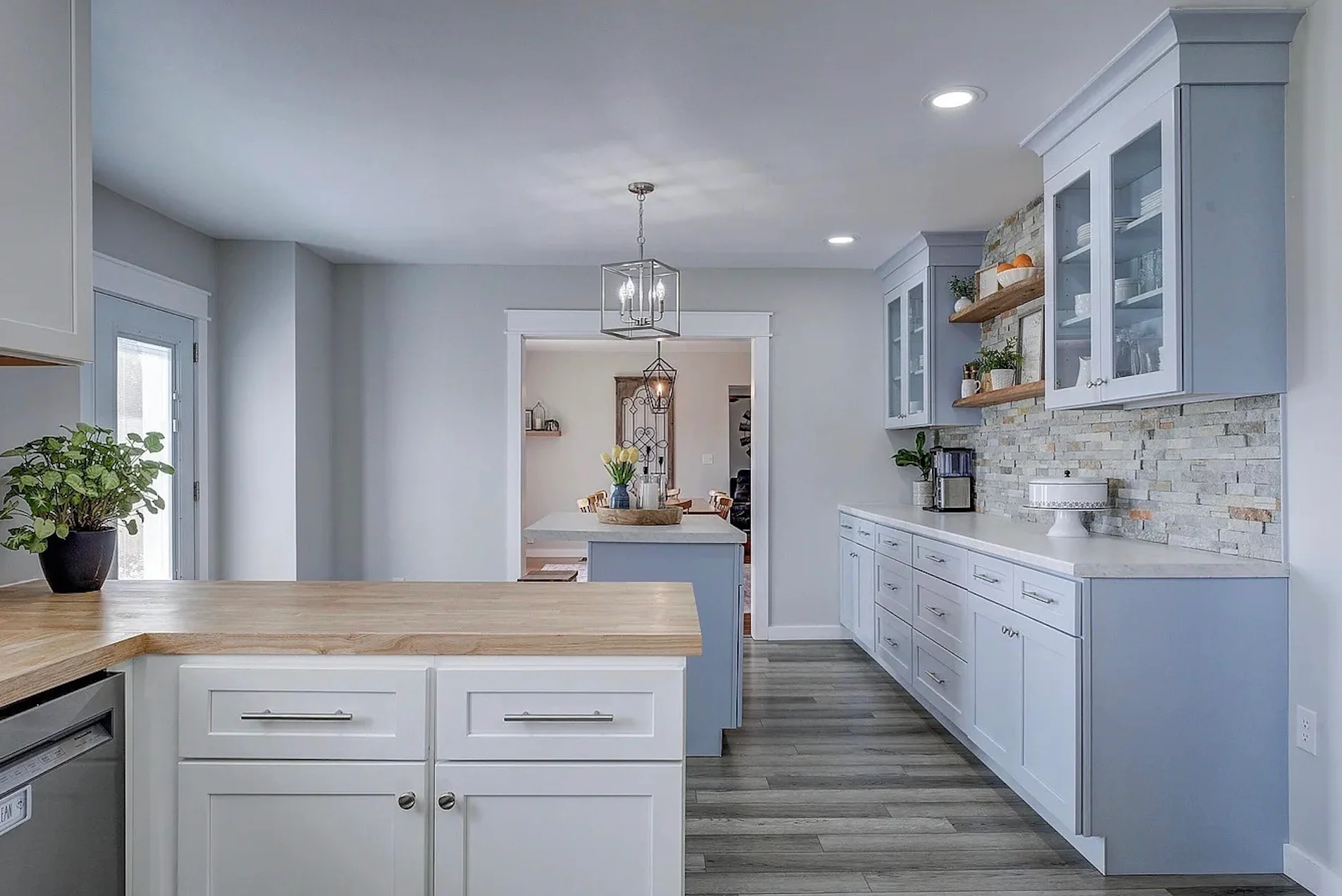 Premade kitchen cabinet layout in a bright modern kitchen with light blue shaker cabinets, a long counter run, glass-front uppers, and a stone tile backsplash.