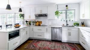 Low price kitchen cabinet idea in a bright U-shaped white shaker kitchen with stainless steel dishwasher, subway tile backsplash, black pendant lights, and a patterned rug.