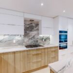 Modern condo kitchen designed by professional kitchen suppliers, featuring handleless white cabinets, wood-tone base units, marble backsplash, and integrated built-in appliances.