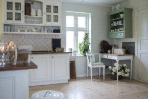 Bright airy kitchen with white shaker-style flat pack kitchen cabinet units, glass-front uppers, wood countertops, and a sage-green hutch near a sunny window.