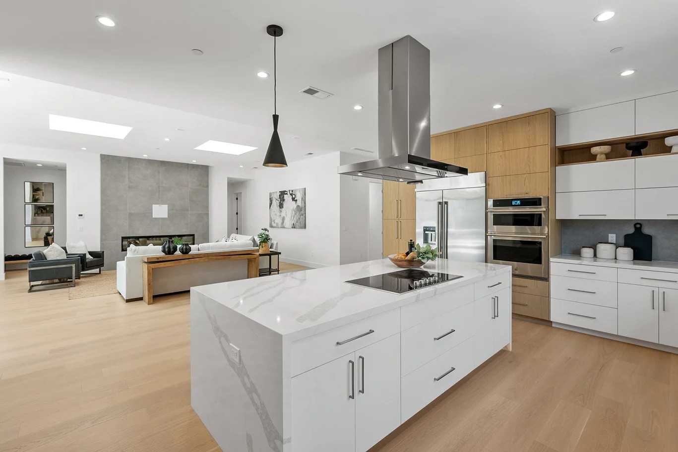 Modern open-concept kitchen with a white waterfall quartz island, two-tone white and light wood cabinetry, and stainless built-in ovens from a kitchen cabinet supplier.