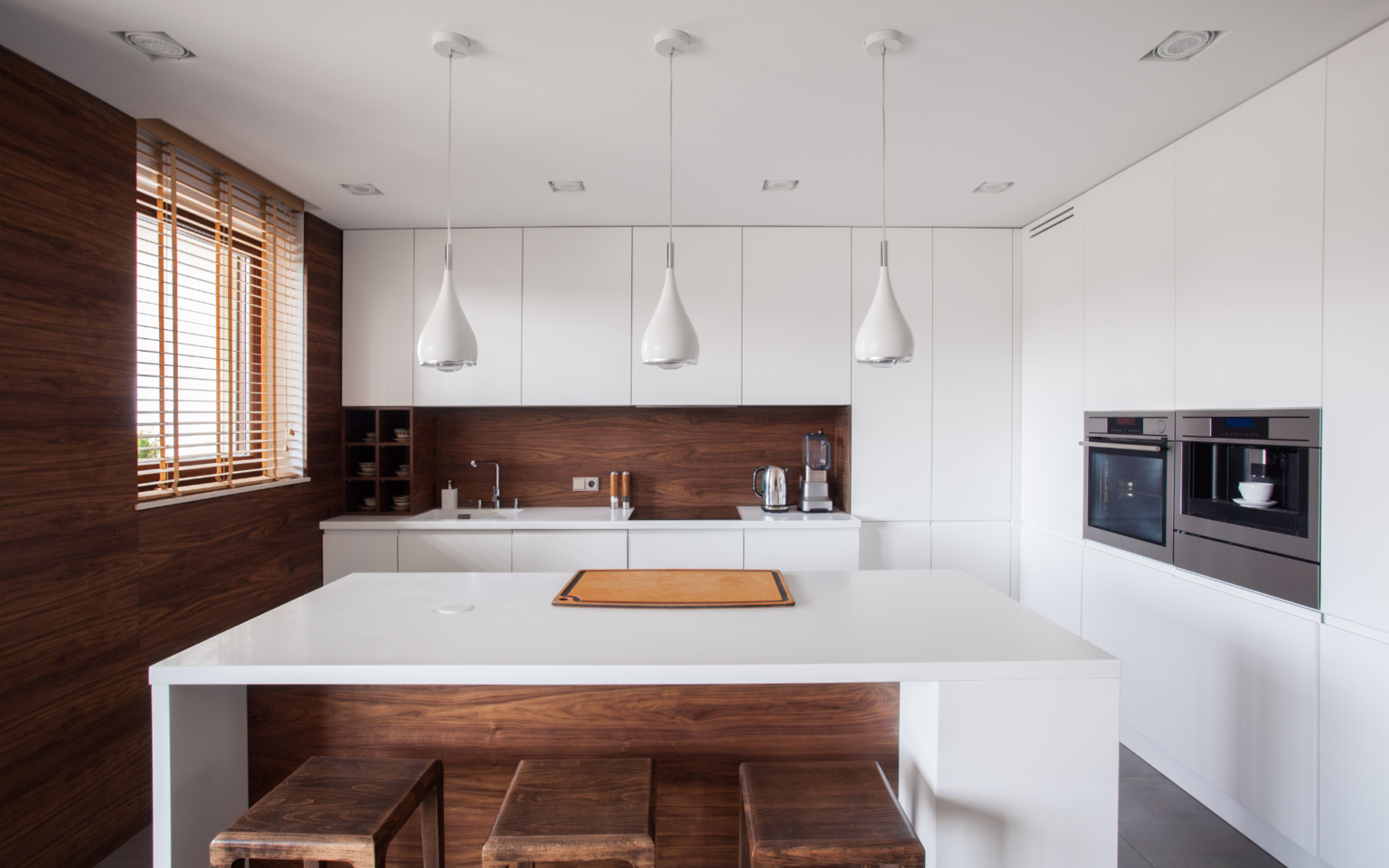 Modern minimalist kitchen featuring white flat pack kitchen cabinet units with warm wood wall panels, a large island, and three pendant lights.