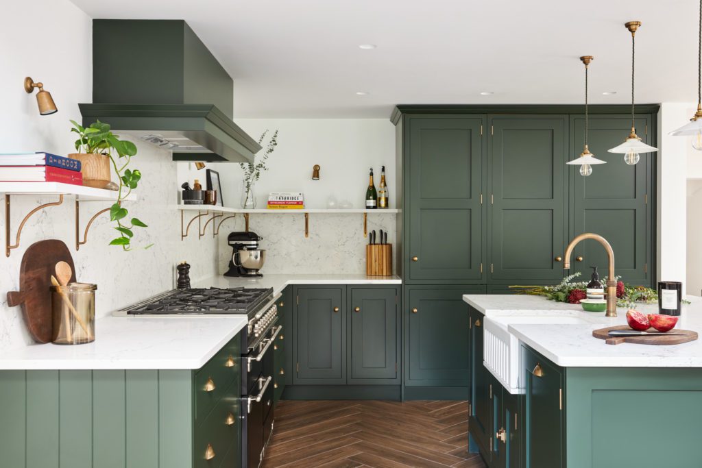 Kitchen suppliers showcase a modern kitchen with dark green Shaker cabinets, brass hardware, white stone countertops, open shelving, and a farmhouse sink.