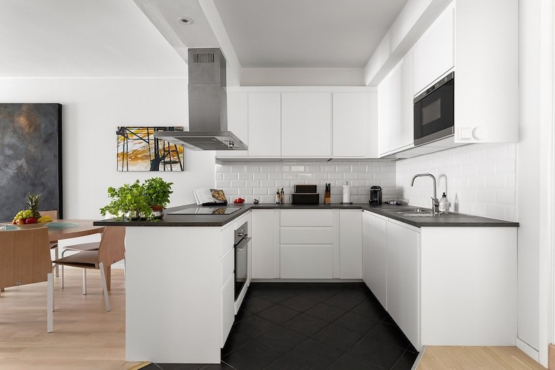 Modern white flat pack kitchen cabinet with L-shaped layout, minimalist design, integrated appliances, and black countertop in a contemporary open-concept home