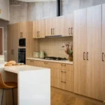 Premade kitchen cabinet set in a modern minimalist kitchen with light oak wood finish, black pulls, tall pantry storage, and a white waterfall island.