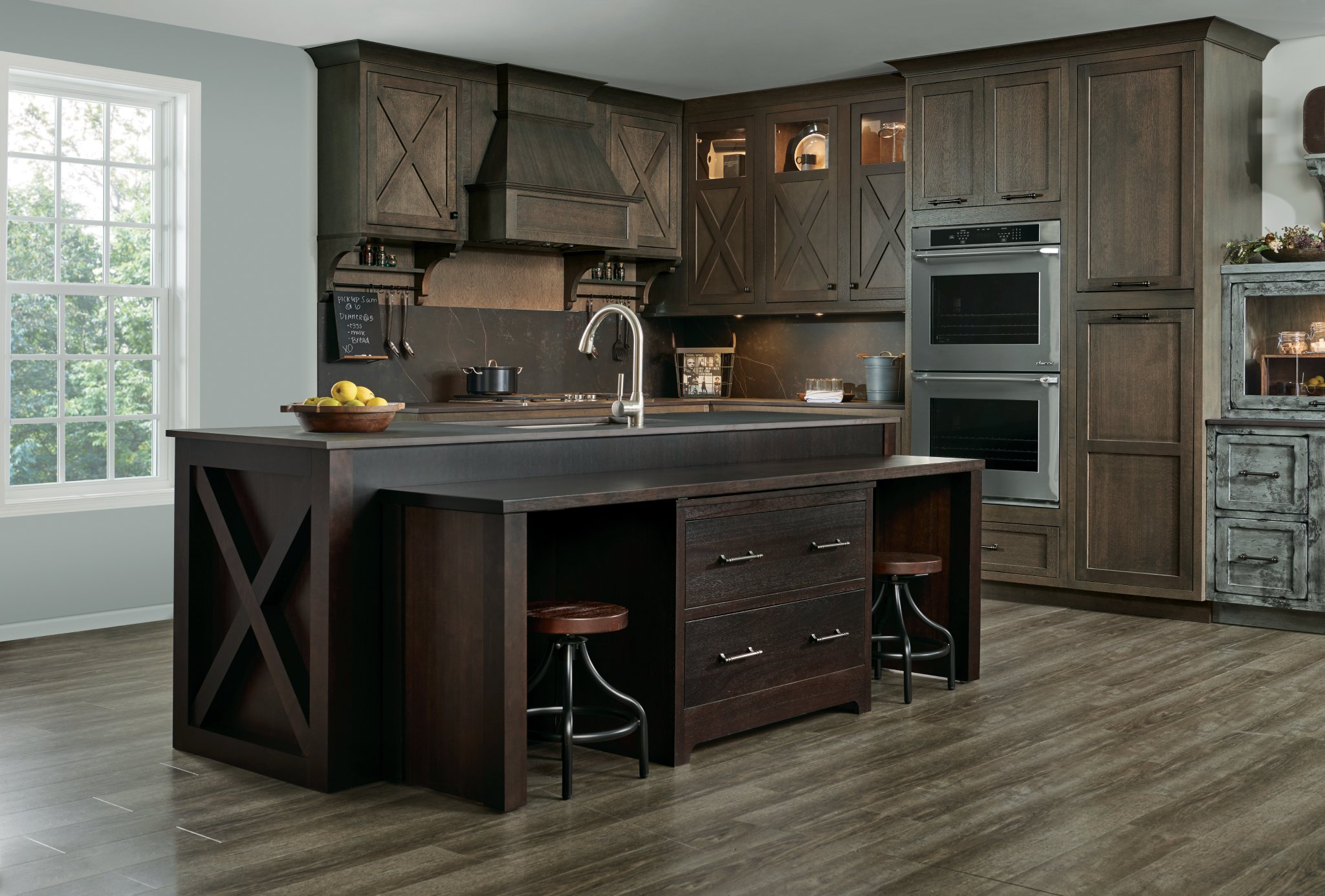 Dark wood premade kitchen cabinet set with X-panel doors, glass uppers, built-in double oven, and a large island with seating.