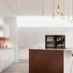 Modern white flat-panel kitchen cabinets with a large island, pendant lighting, and built-in wall ovens in a bright contemporary kitchen.