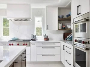 Modern white premade kitchen cabinet set with black pulls, quartz countertops, and built-in wall ovens in a bright contemporary kitchen.